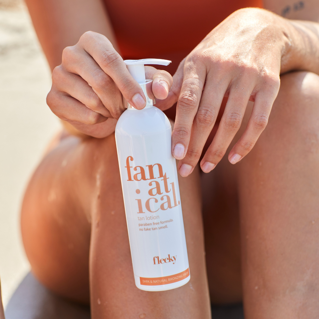 Model sitzt im Sand und hält die sanfte Fleeky Self Tan Lotion in der Hand